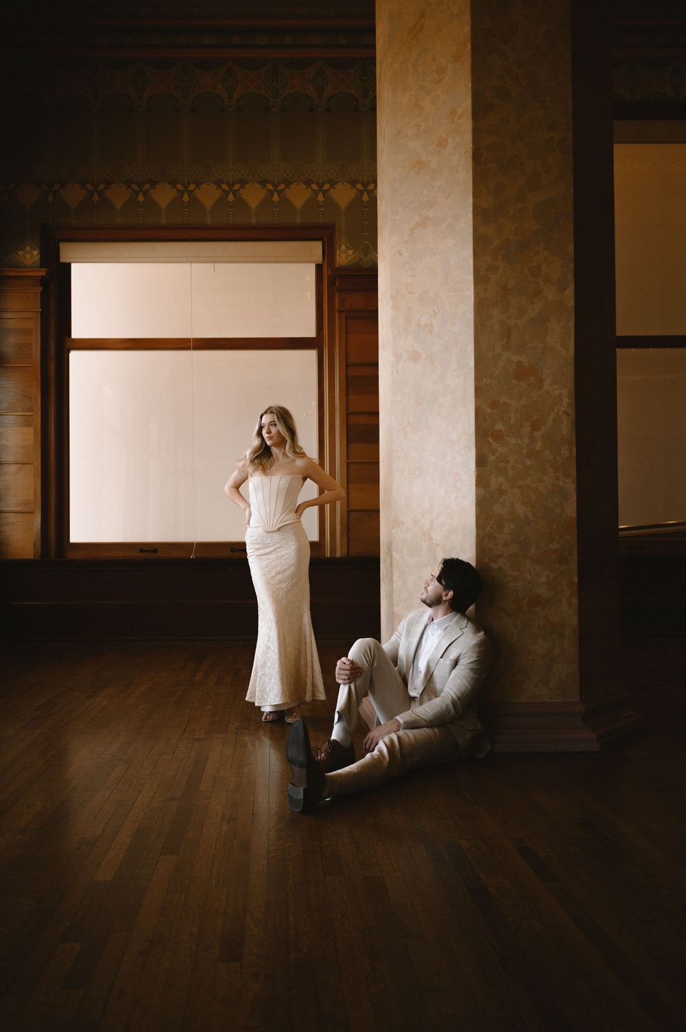 Bride in white gown stands as groom sits against column on hardwood floor in vintage ballroom.