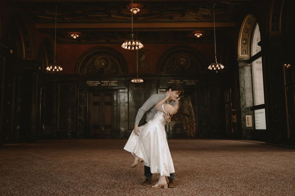 Couple sharing a romantic dip kiss in an elegant grand hall with ornate architecture and warm chandelier lighting.