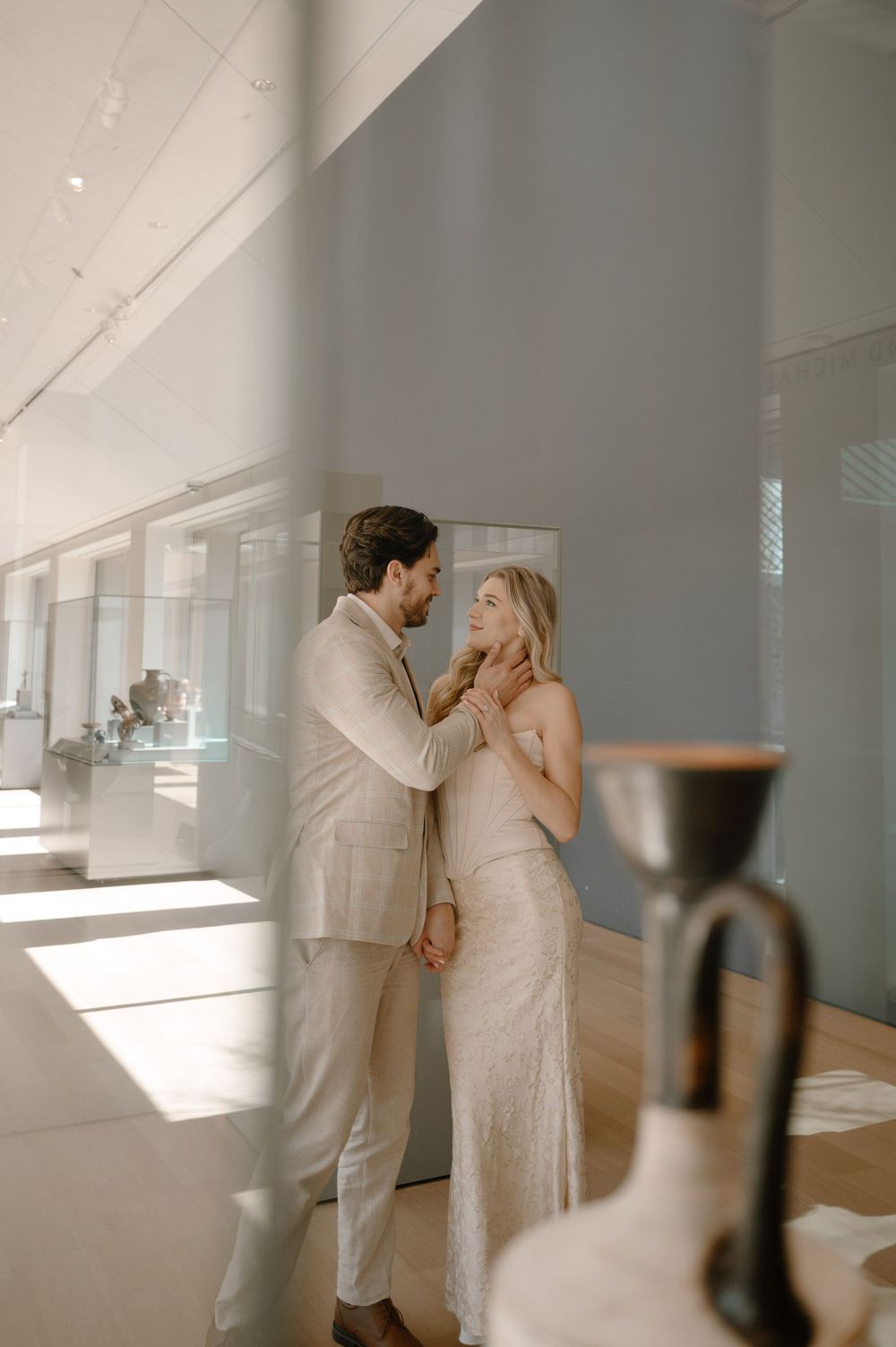 Couple sharing a tender moment in a sunlit museum gallery, dressed in elegant neutral attire.