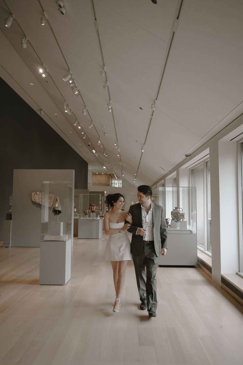 A couple walks through a modern art museum gallery, the woman in a white dress and the man in a suit.
