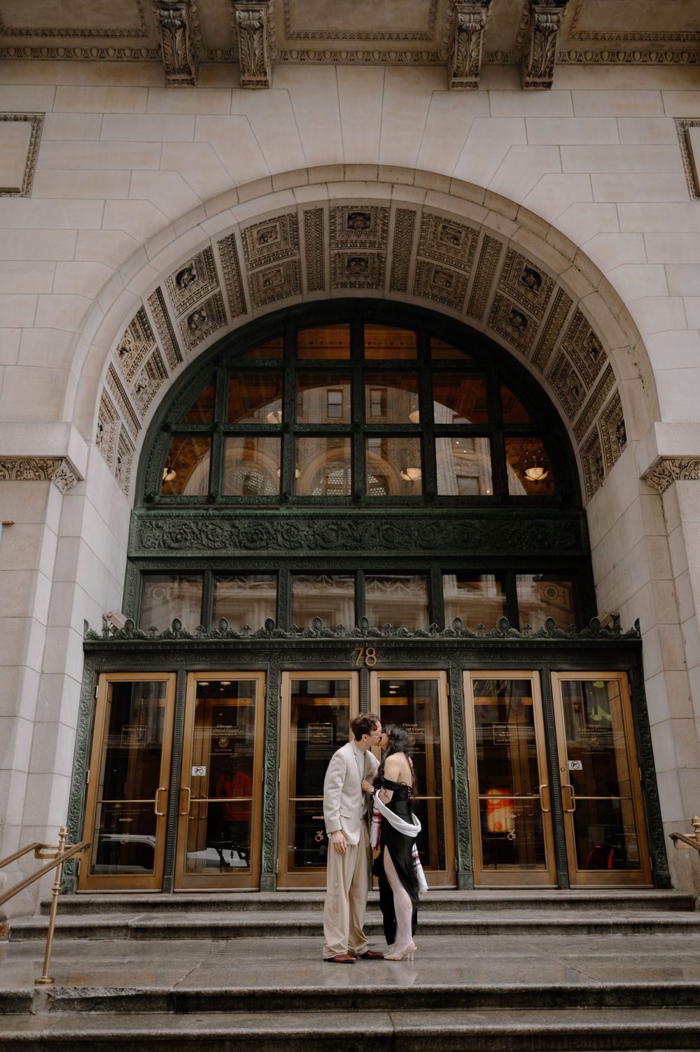 Couple kissing under ornate arched entrance of a classic beaux-arts building exterior.