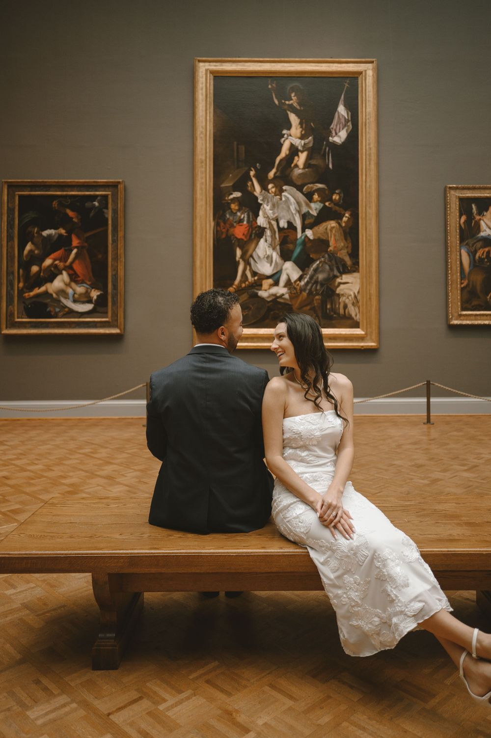Couple in wedding attire sitting on a museum bench admiring classical paintings in an art gallery.