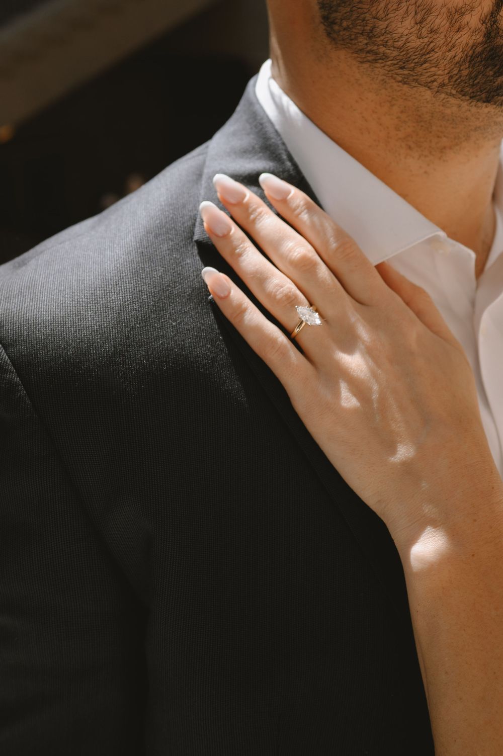 A woman's hand with a diamond engagement ring rests on a man's shoulder in a dark suit.