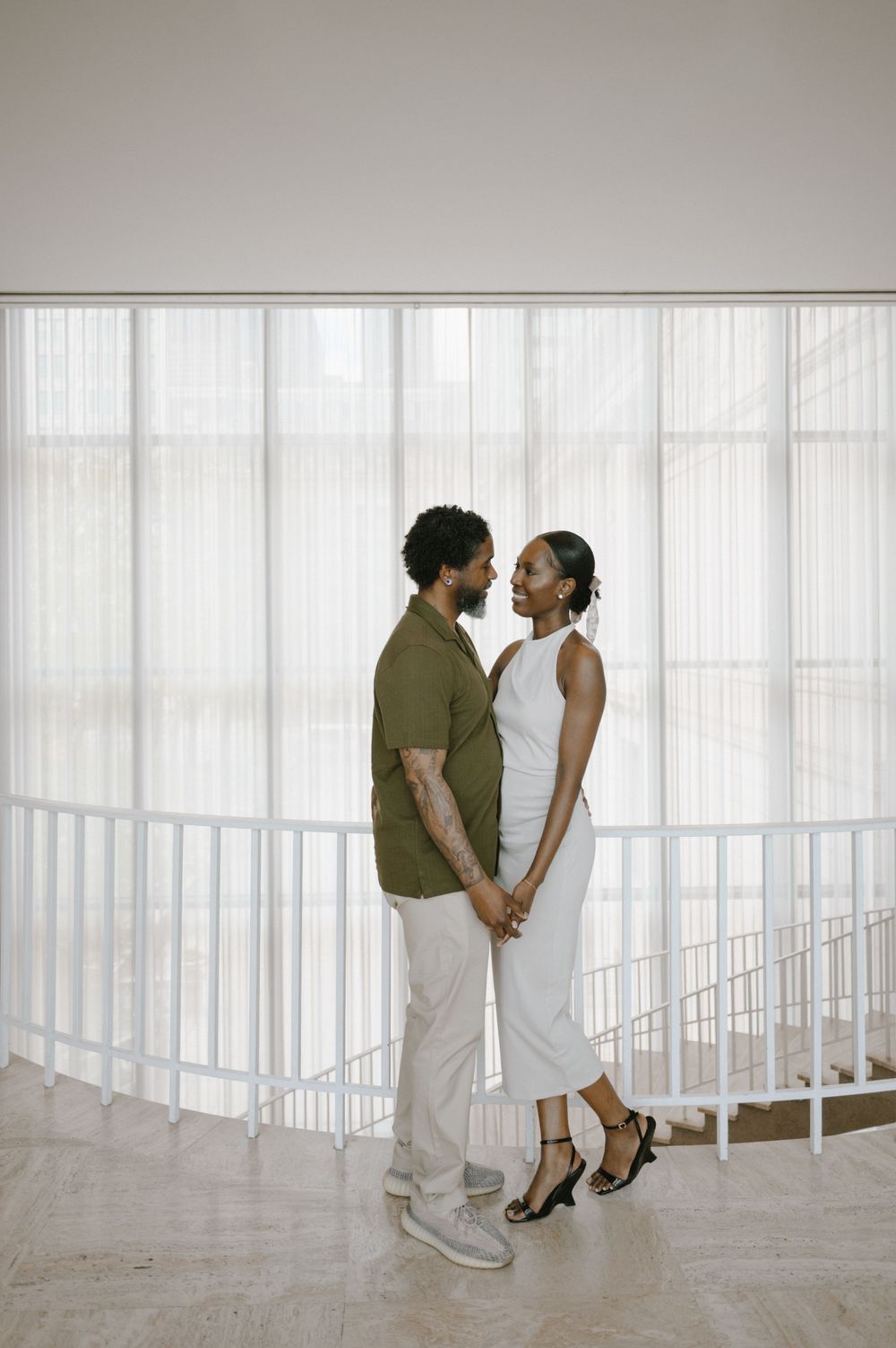 A couple holds hands and gazes at each other in a bright modern space with sheer white curtains.