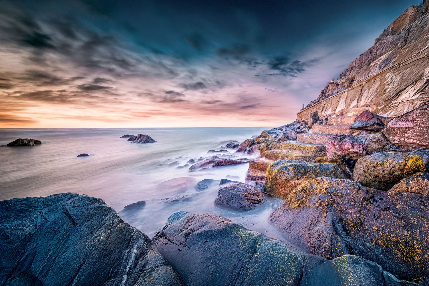 Long exposure landscape of a rugged coastline with colorful sunset clouds reflecting on the ocean waves.