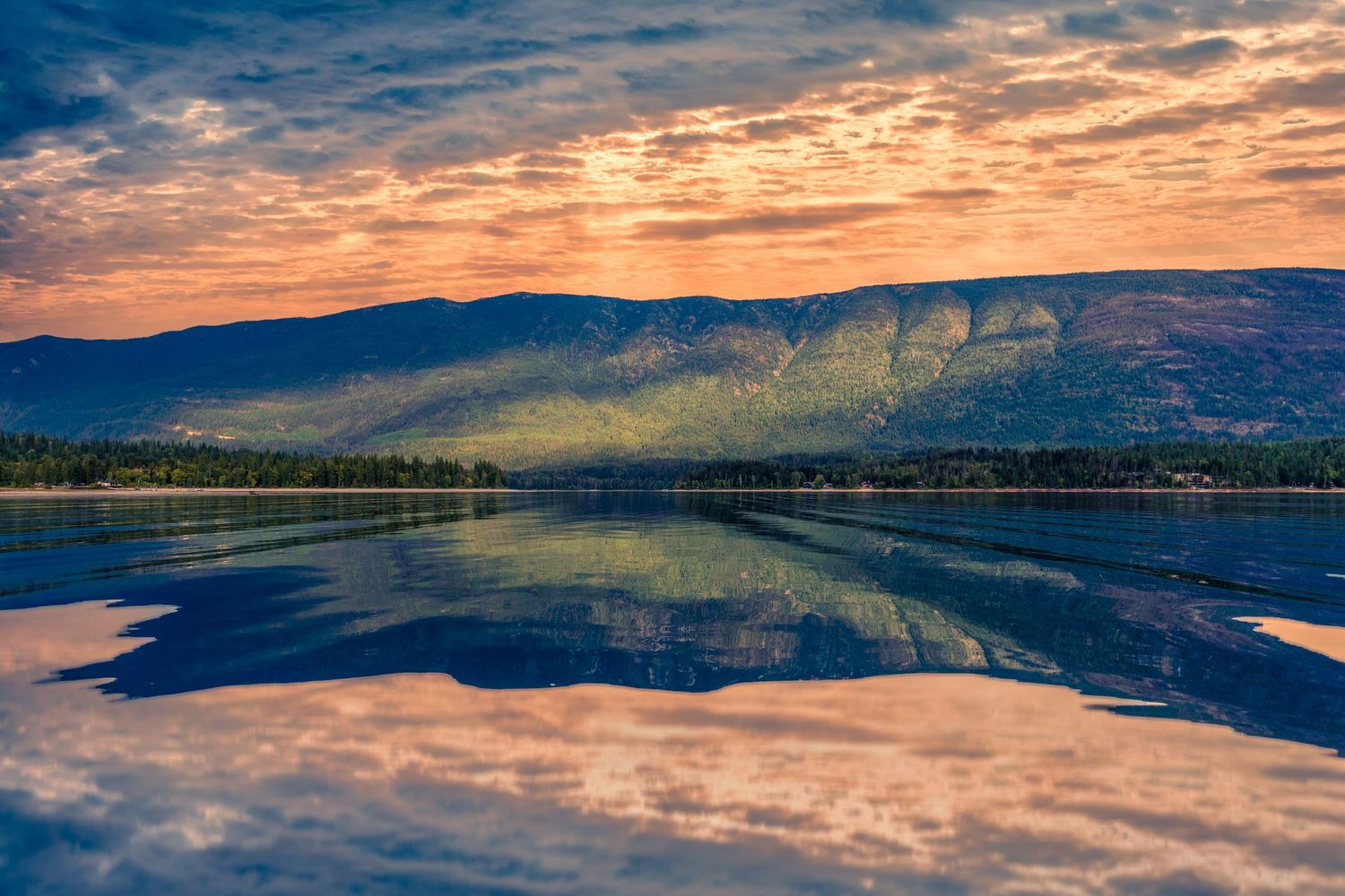 Dramatic sunset sky reflects in calm lake waters with mountains and forests creating a mirror-like landscape.