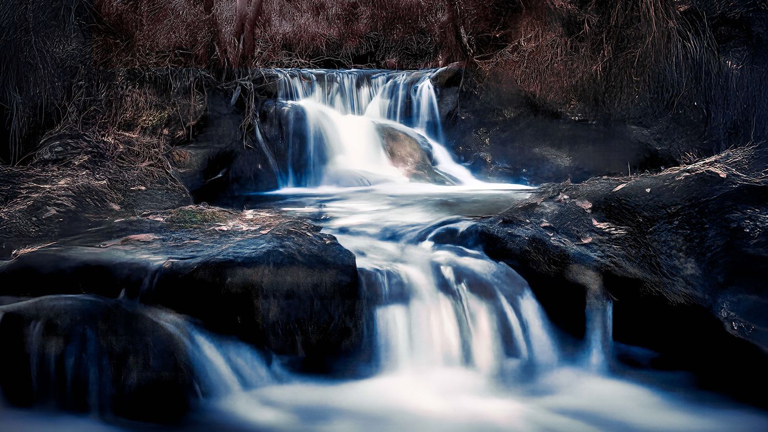 Moody long exposure of a small forest waterfall cascading over dark, mossy rocks.