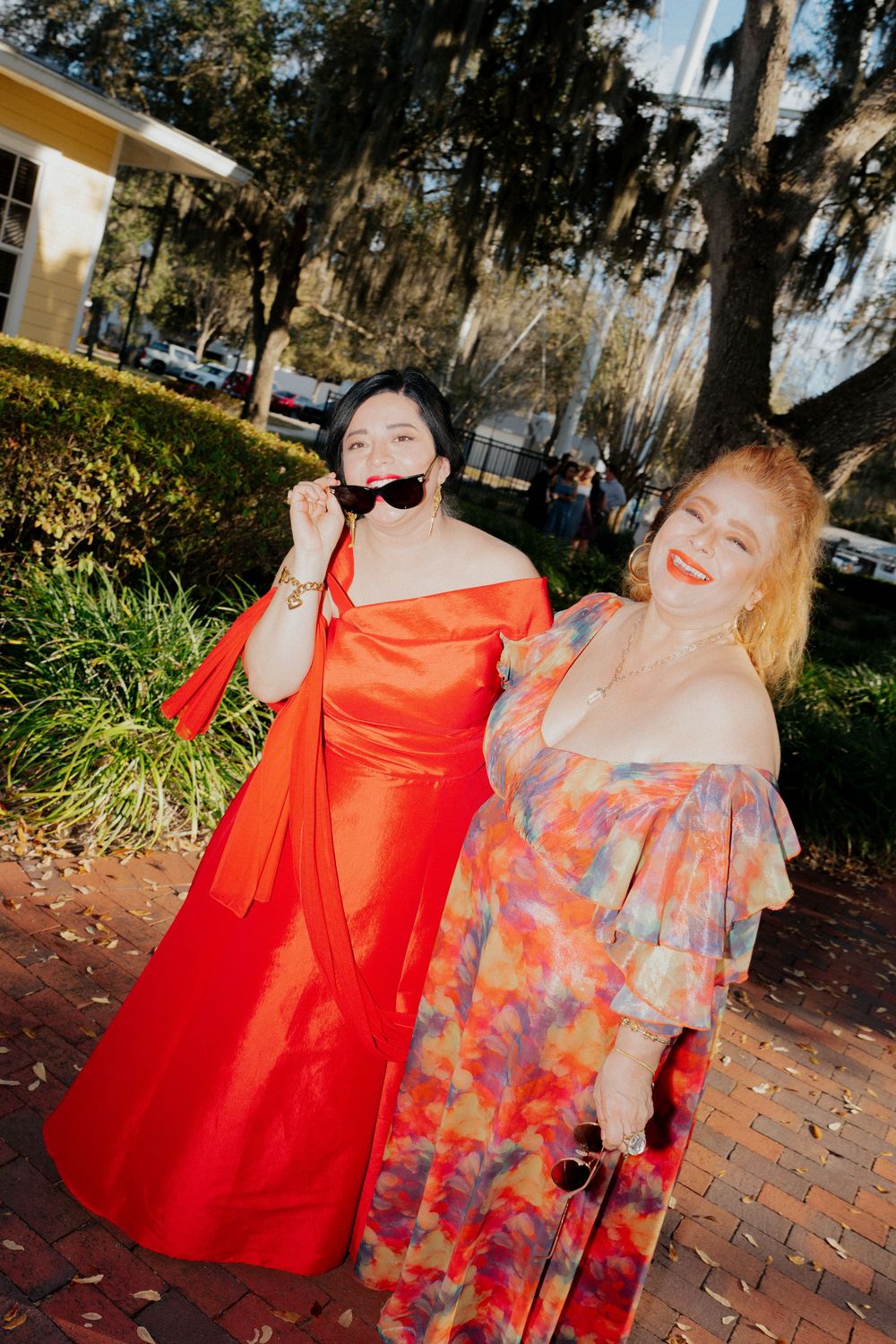 Two women in elegant off-shoulder dresses, one red gown and one floral, posing outdoors on a brick path.