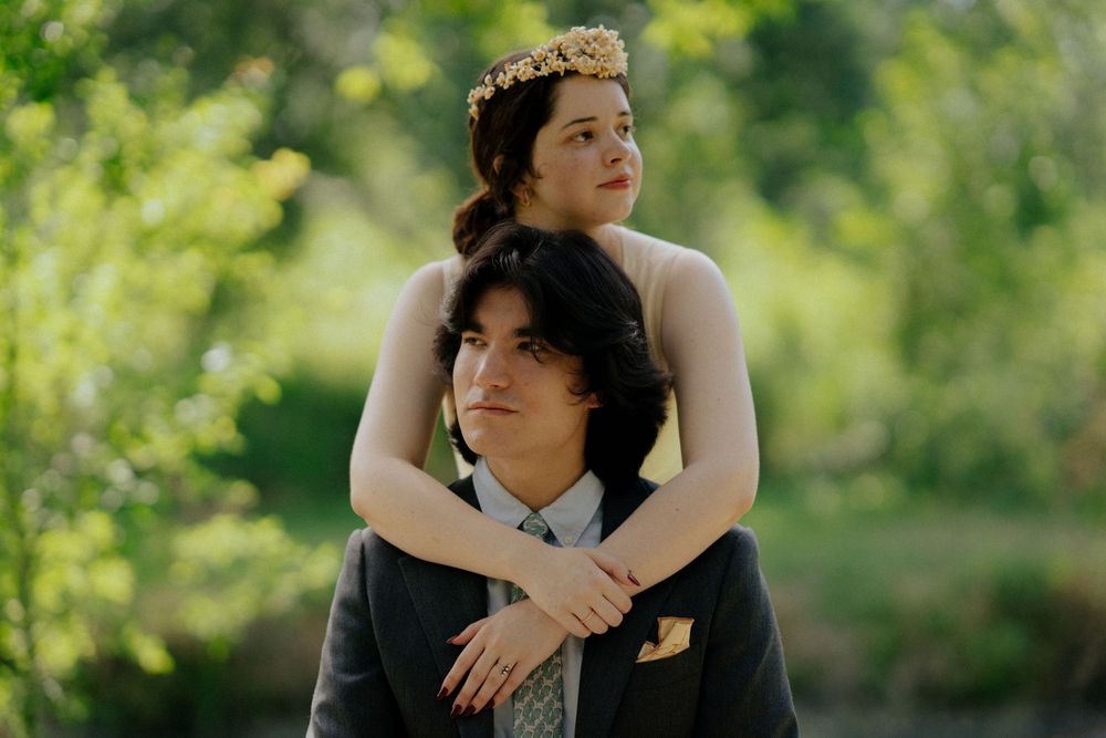 Couple posing outdoors, woman with floral crown resting on man's shoulders in a lush green garden setting.