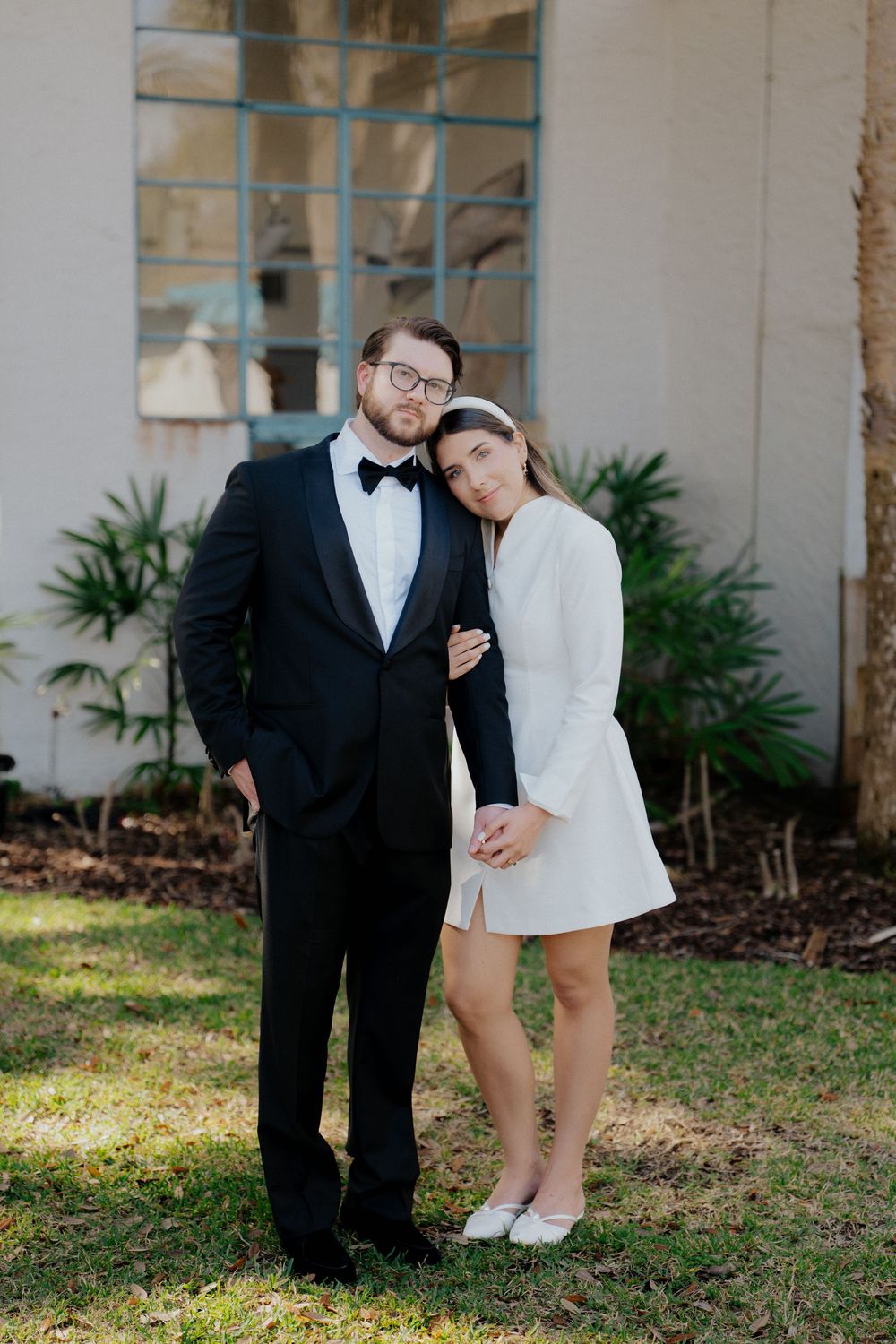 Couple in wedding attire, man in black tuxedo and woman in white mini dress, posing outdoors near a building.