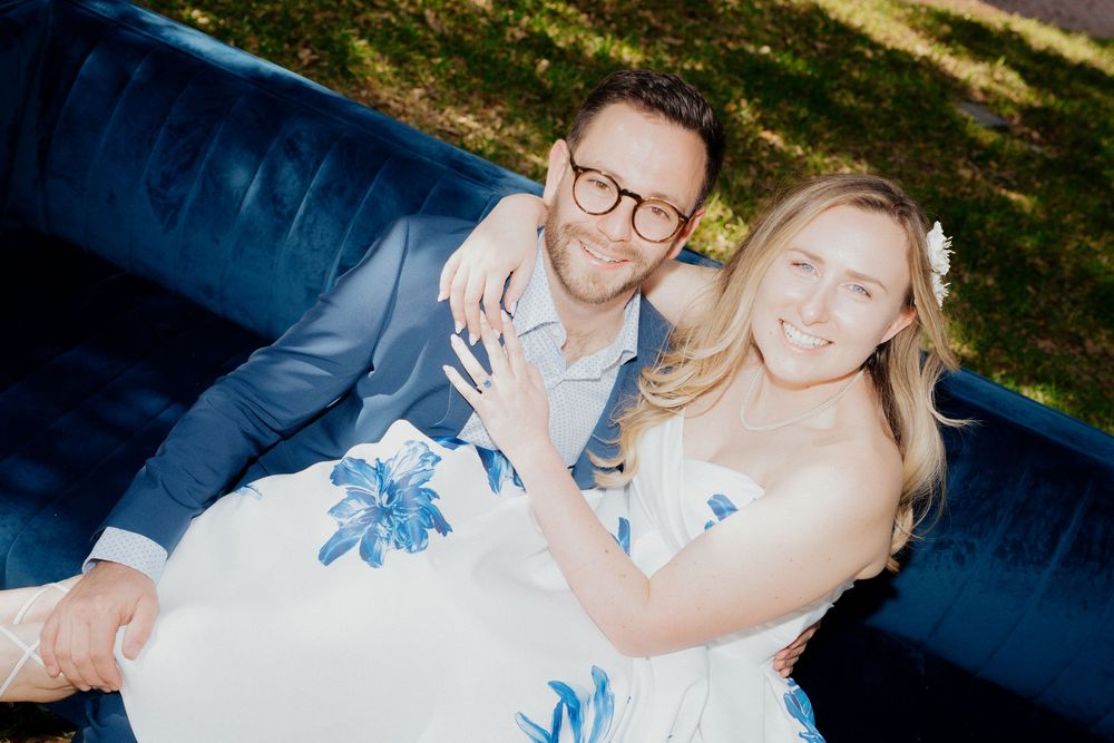 Happy couple sitting on a blue velvet couch outdoors, woman in floral dress, man in blue suit, smiling.
