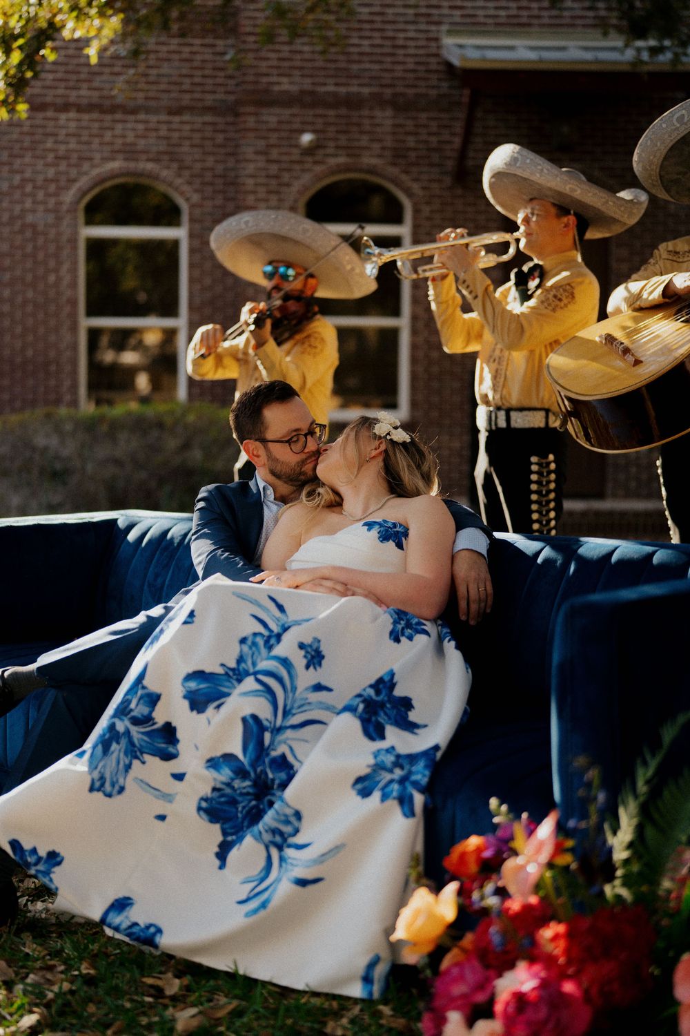 Couple kissing on blue sofa with mariachi band in sombreros playing trumpet at outdoor wedding celebration.