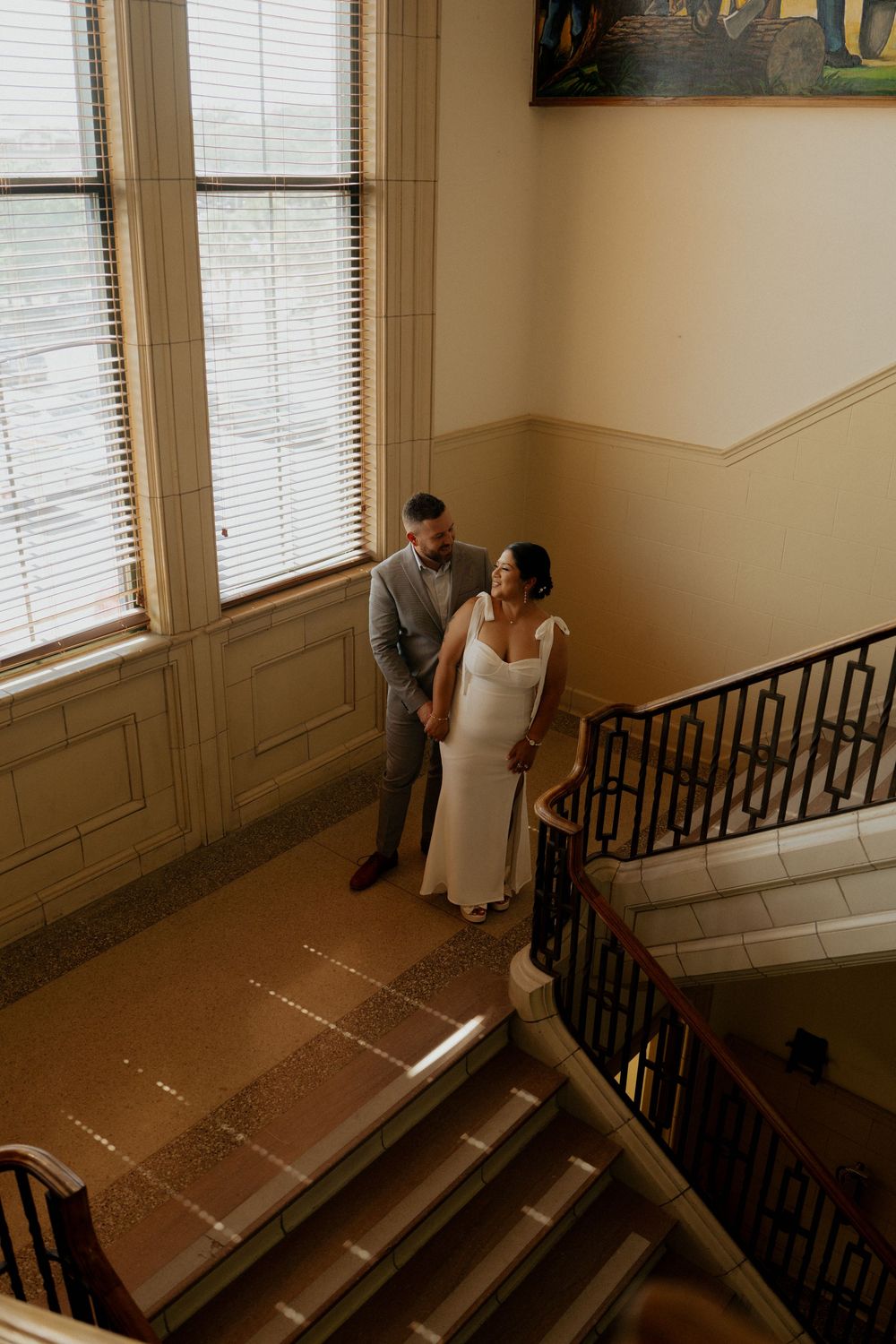 Bride and groom stand together on a sunlit staircase in an elegant venue, captured from above.