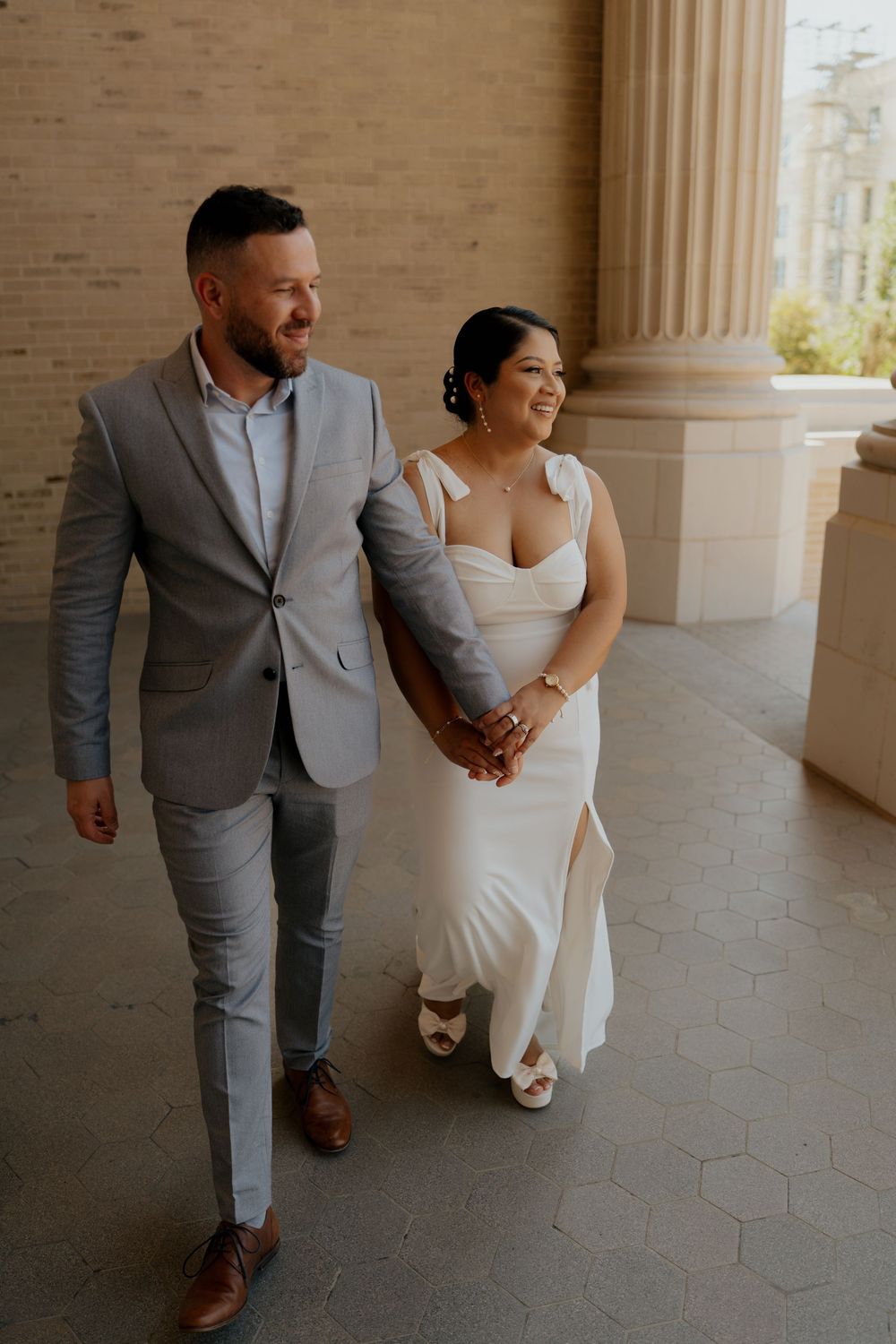 A smiling bride in a white gown and groom in a gray suit walk hand in hand through an elegant columned venue.