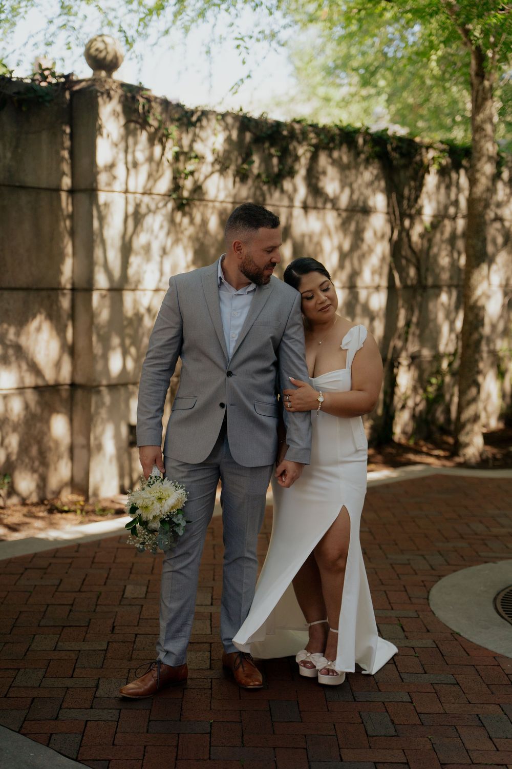 Bride in white gown and groom in gray suit hold flowers on brick path with ivy wall backdrop.