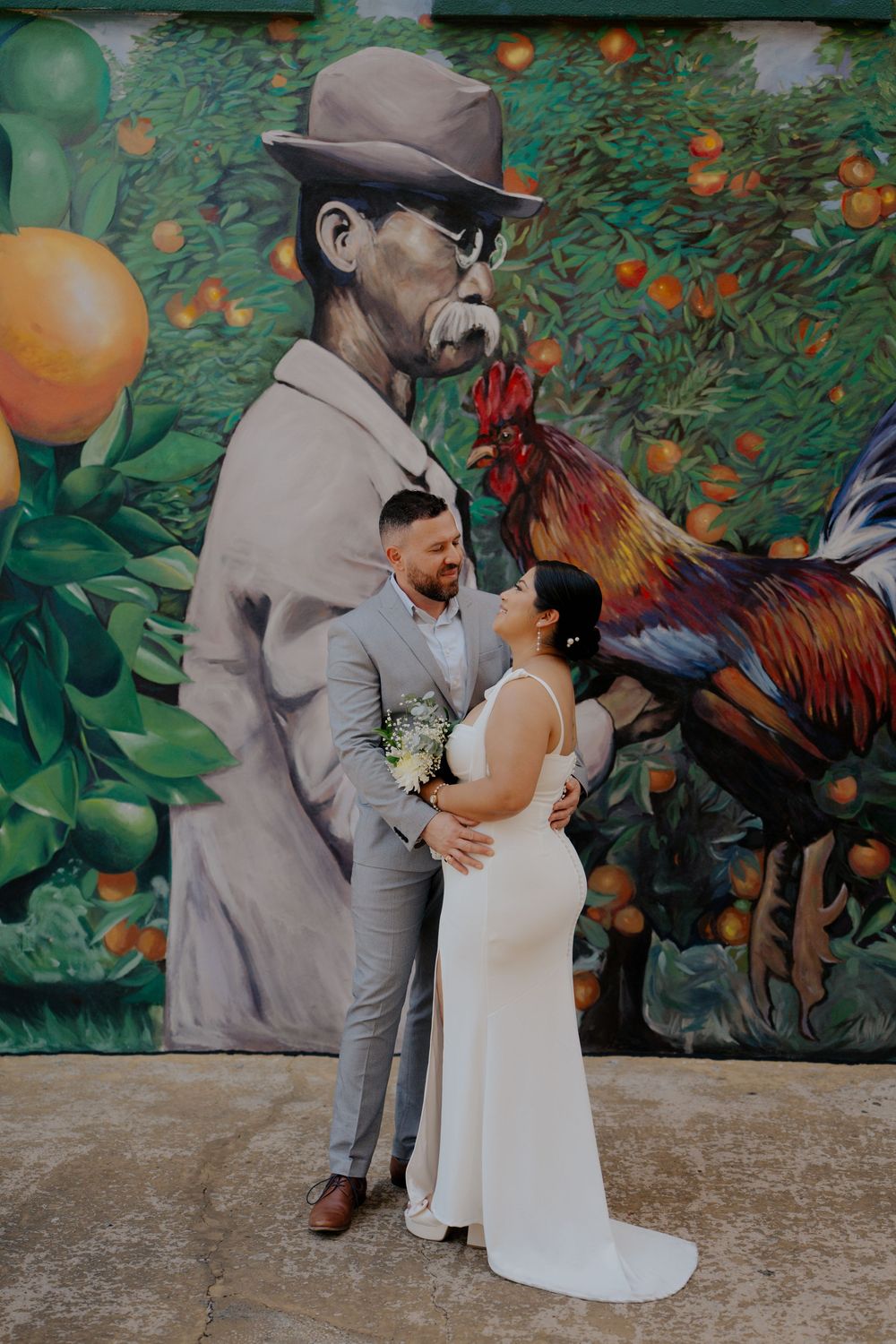 Bride and groom pose in front of a vibrant rooster and farmer mural with orange trees on their wedding day.