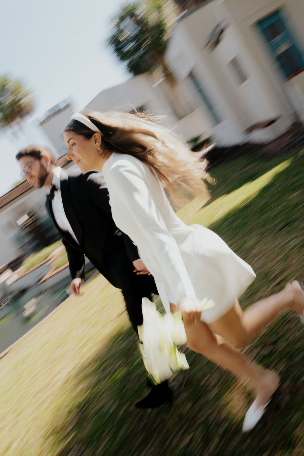 Bride in white mini dress runs with groom in black suit across green lawn, motion blur capturing joyful wedding moment.