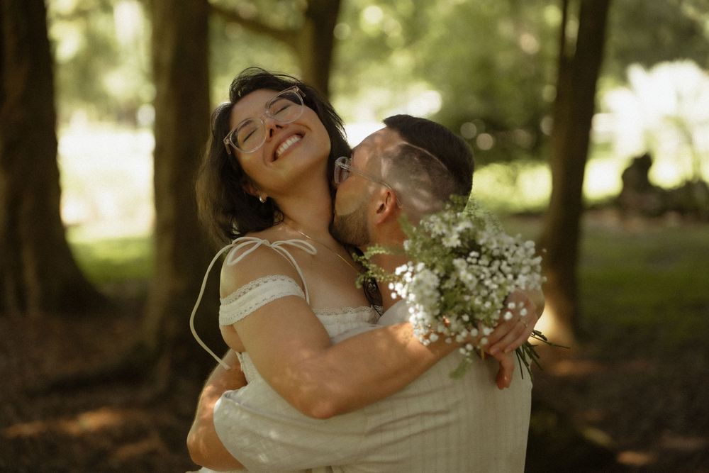 A groom kisses his smiling bride holding a white bouquet in a sunlit forest wedding photo.