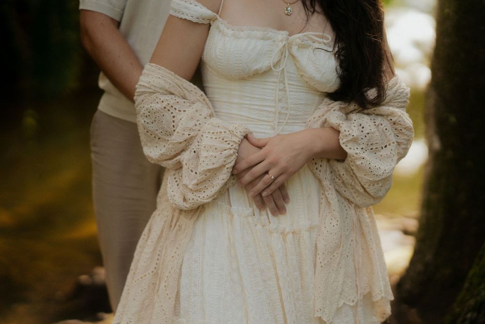 Couple embracing outdoors, woman wearing elegant cream lace Victorian-style dress with puffed sleeves in a forest setting.