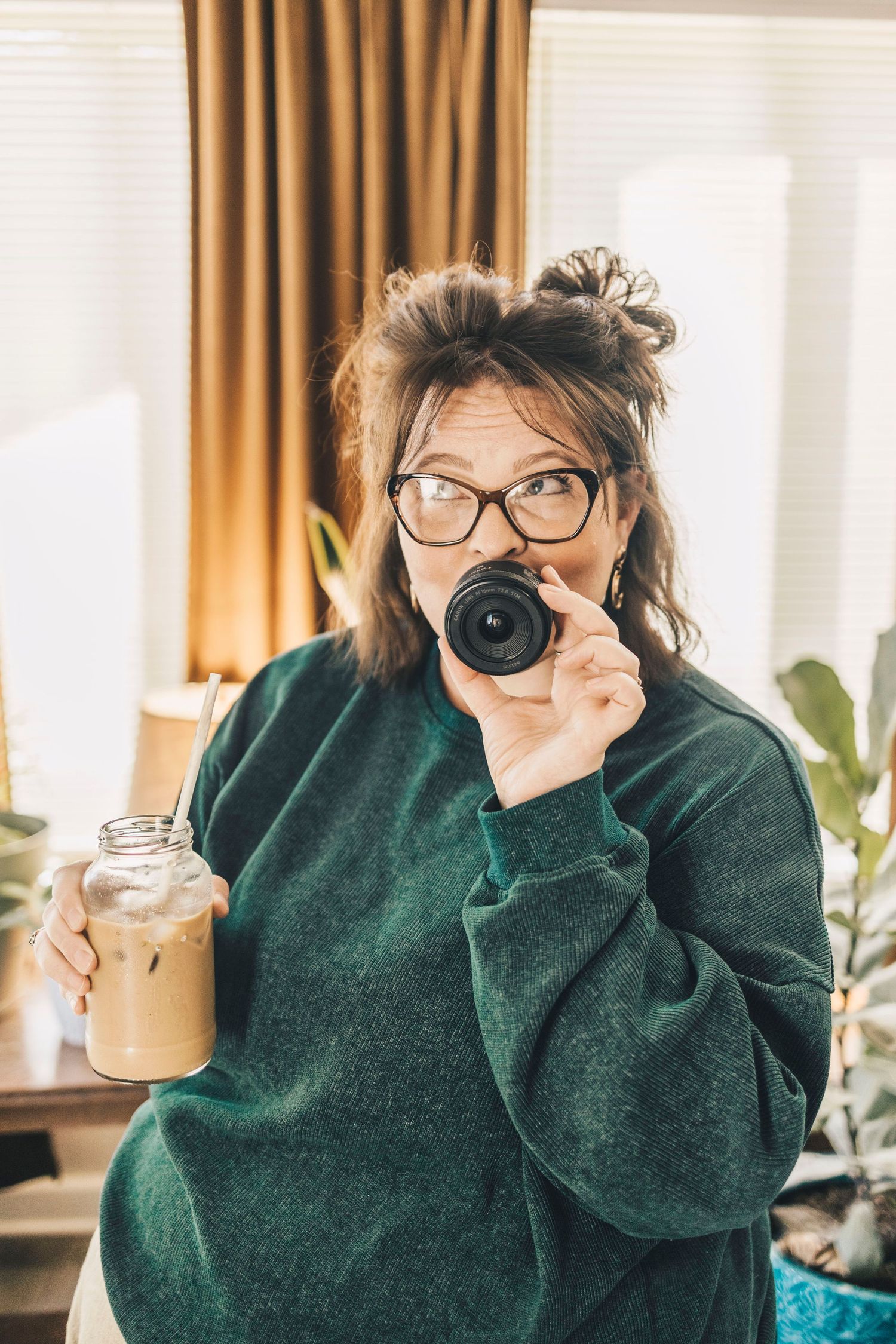 Woman in green sweater holding iced coffee jar while looking through camera lens indoors.