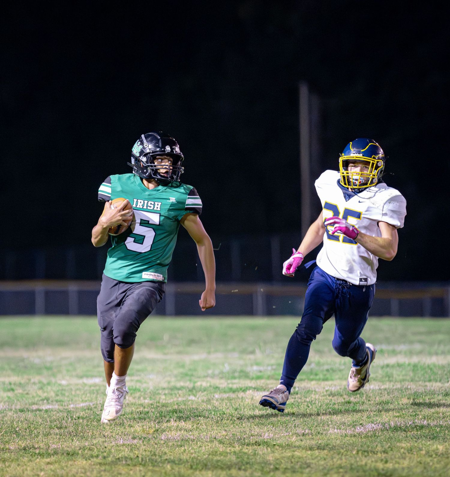 High school football player in green jersey runs with the ball as defender in navy uniform gives chase during night game.