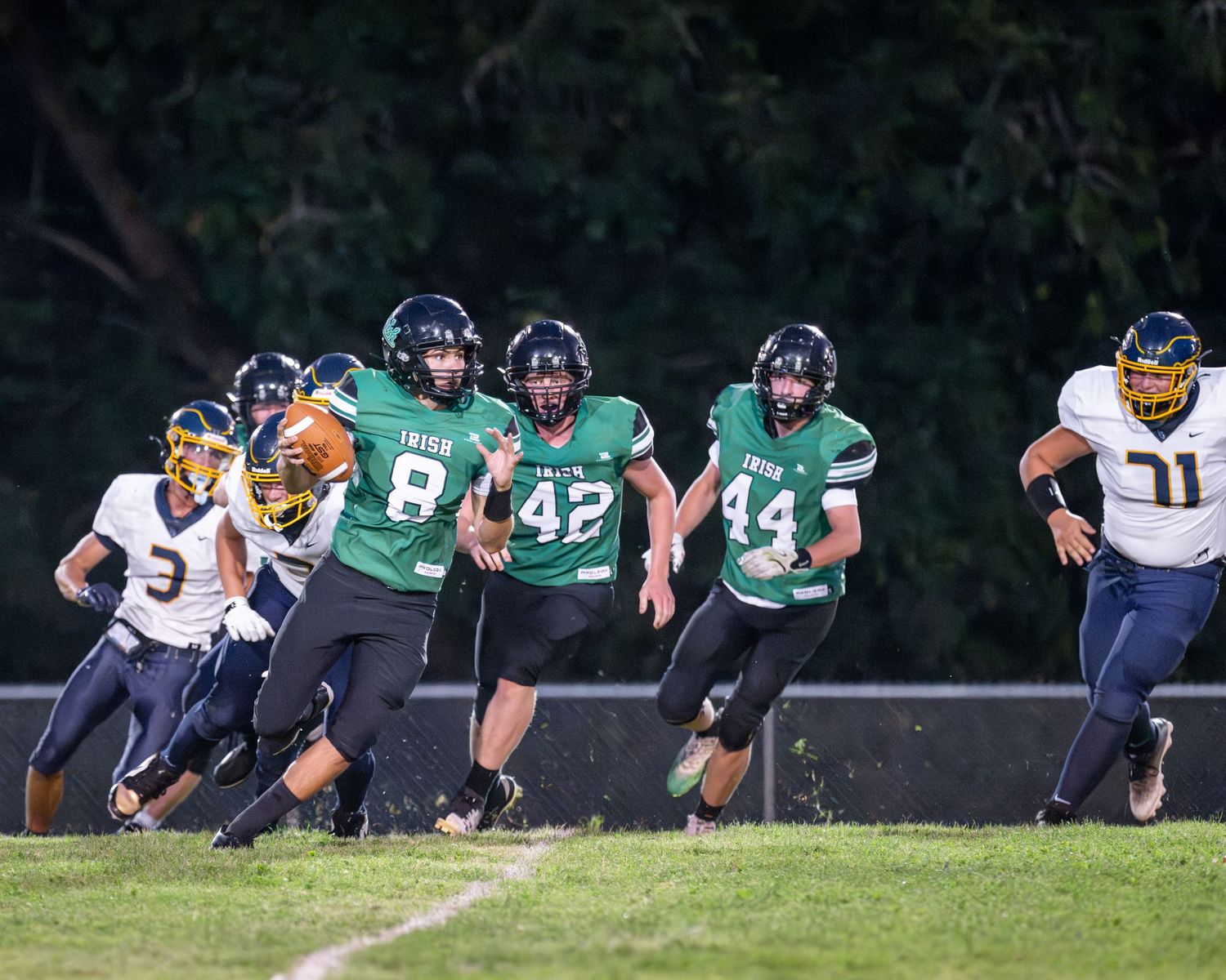 Football team players arranged in a line on grass field wearing green and navy uniforms during practice drill.