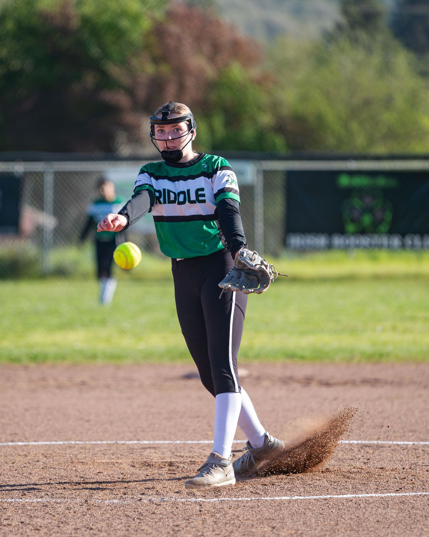 Softball pitcher in green and white uniform delivering pitch on dirt mound.