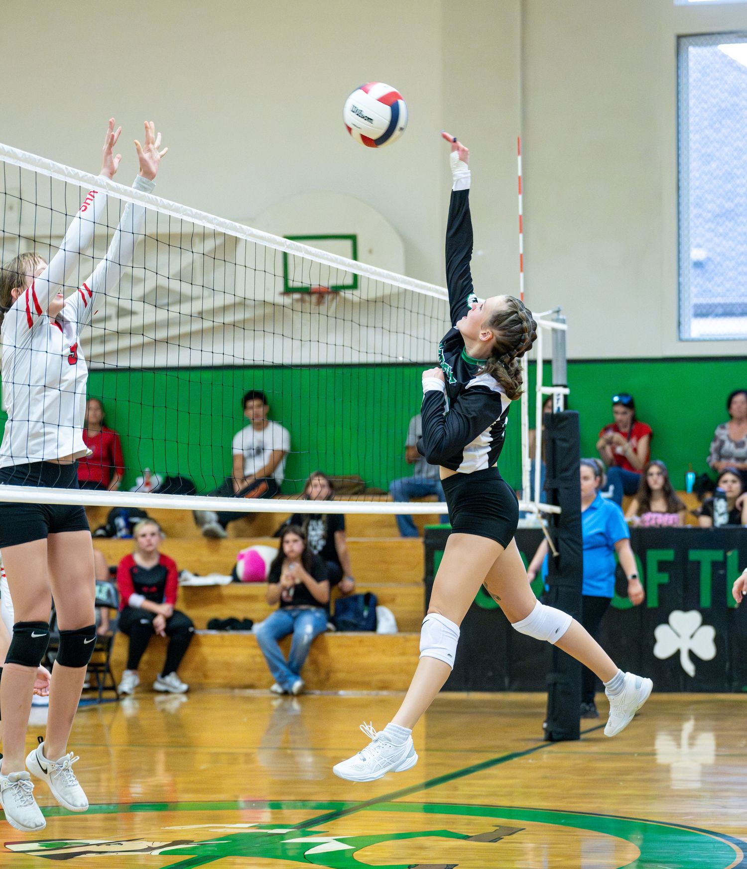 Indoor volleyball players leap to block and spike during an intense game at a gymnasium with green walls.