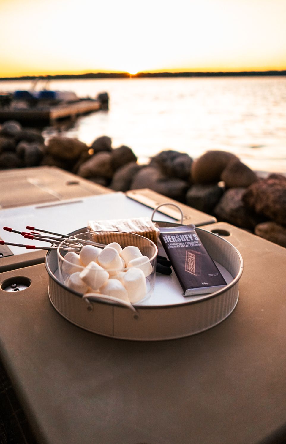 Plate of marshmallows and chocolate bars set up for making s'mores by the water at sunset.