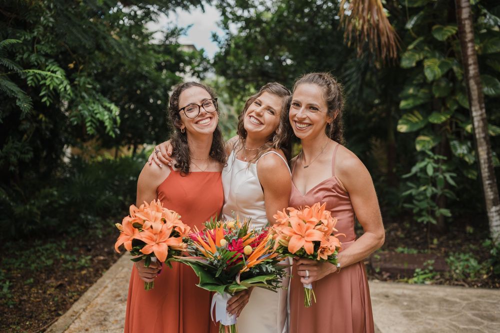 Three women hold vibrant orange and pink tropical flower bouquets in a garden setting, Sandos Caracol Wedding.