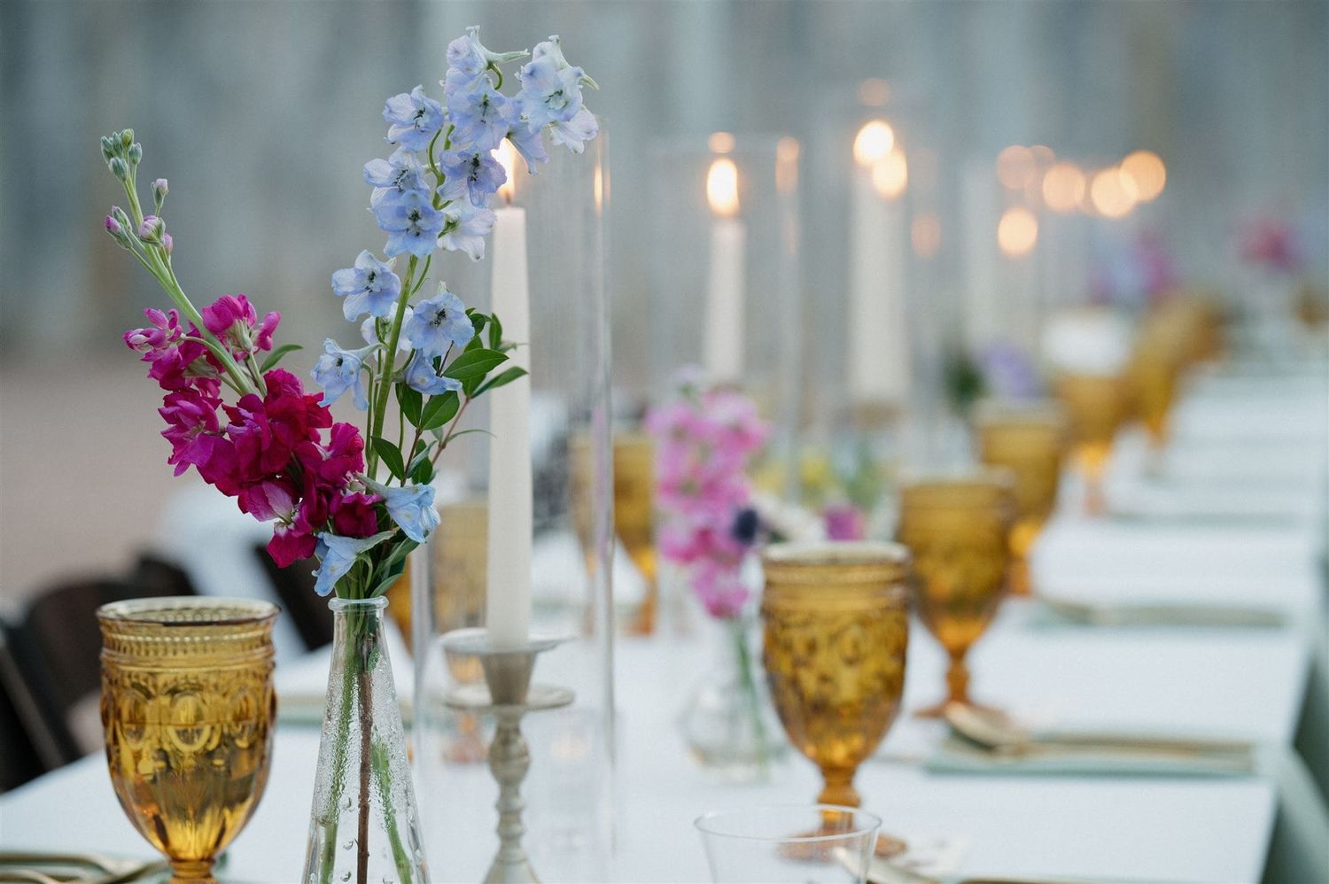 Elegant table setting with amber goblets, white candles, and delicate purple and blue flower arrangements.