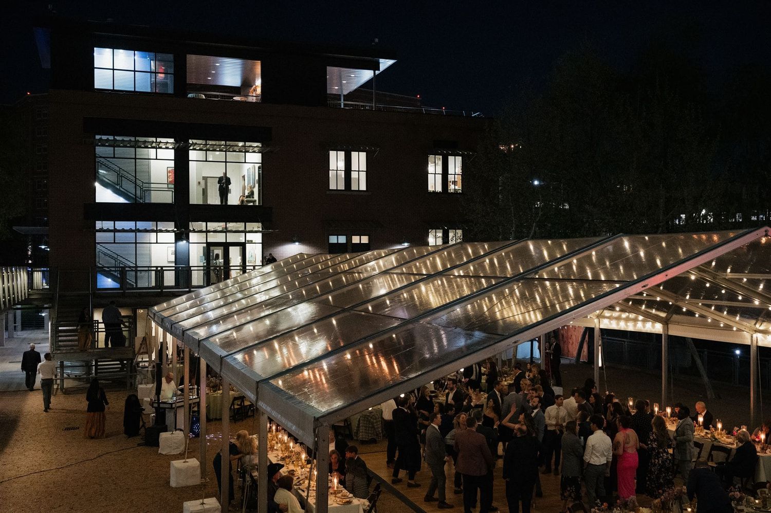 Crowded evening market with illuminated glass roof stalls and modern brick building in background at night.