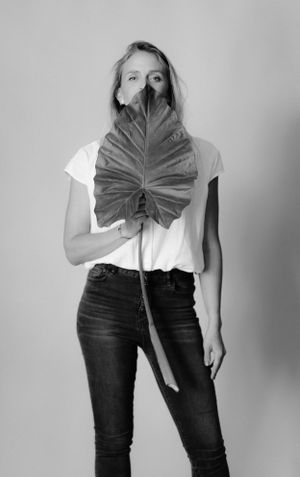 Person holding large tropical leaf in front of face while wearing white shirt and dark jeans.