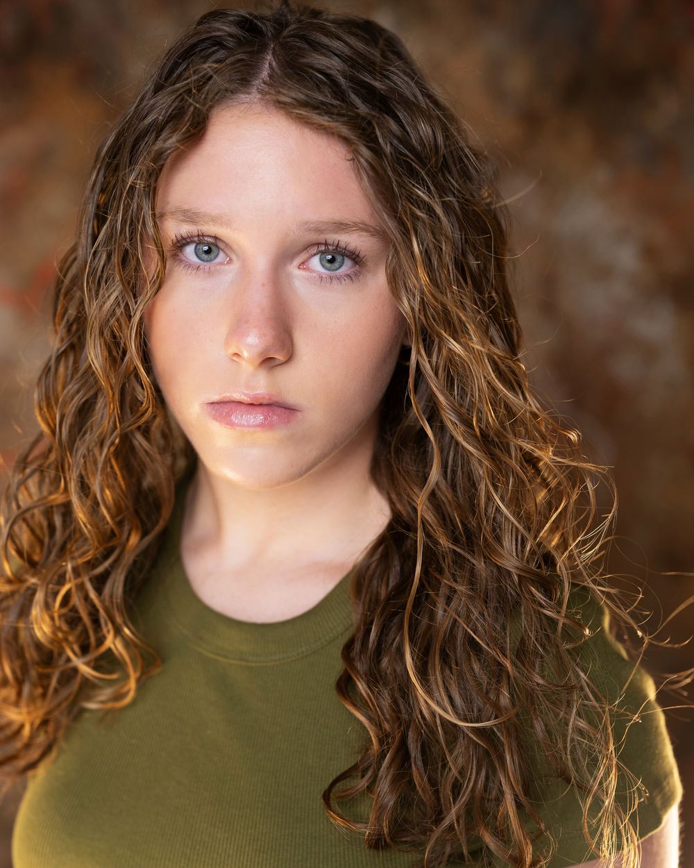 Young woman with long curly brown hair and blue eyes wearing an olive green top, professional headshot photo.