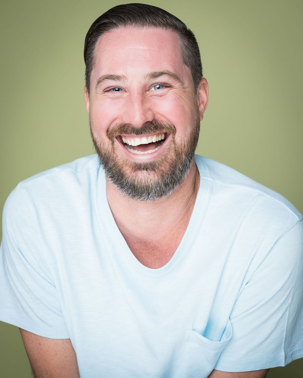 Portrait of a beaming person in a white t-shirt with a full beard against a green background.