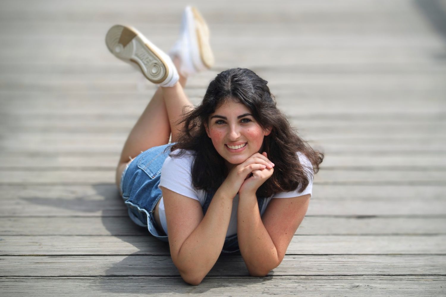Young woman in denim shorts and white top lying on wooden deck, smiling cheerfully at camera.