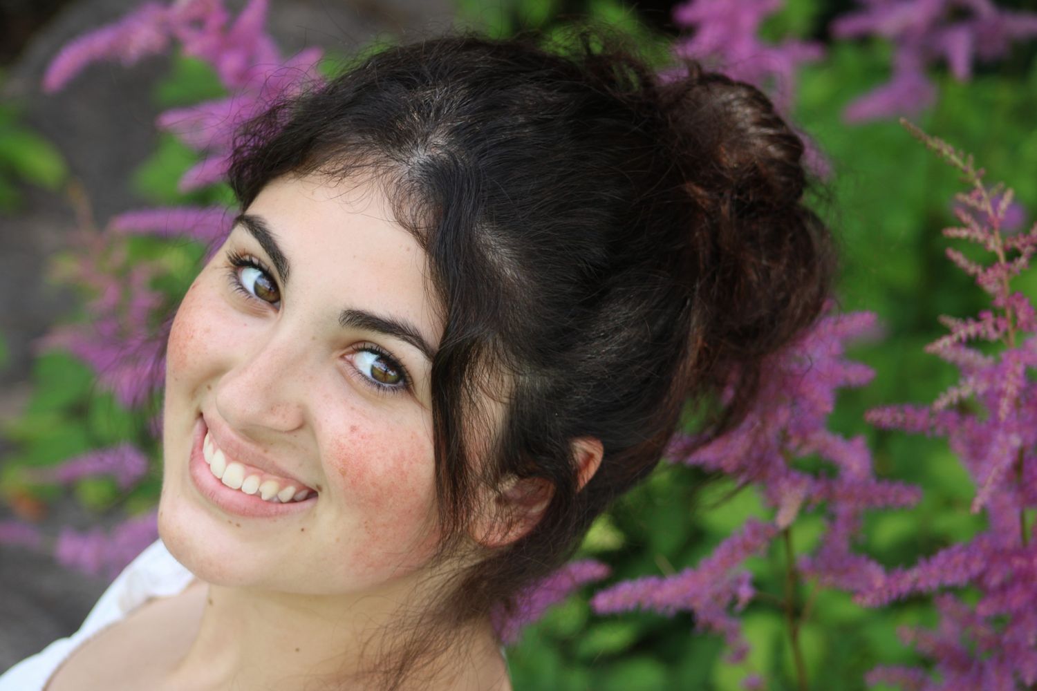 Young woman with dark hair smiling outdoors surrounded by pink astilbe flowers in a garden setting.