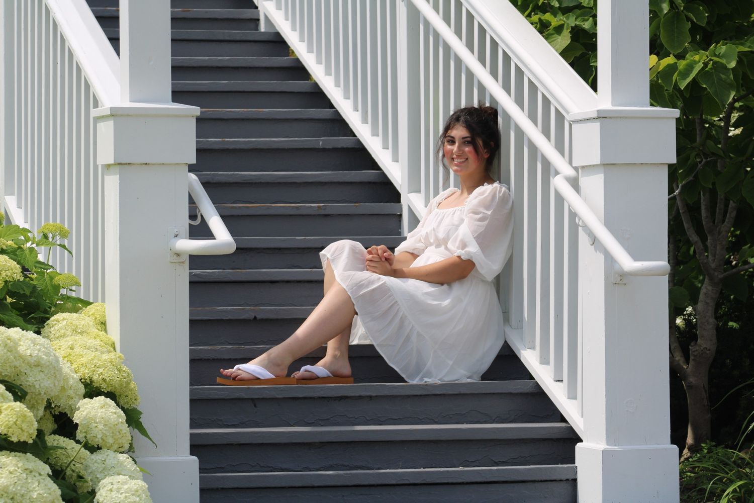 Woman in white dress sitting on gray porch steps beside white railings and blooming hydrangeas.