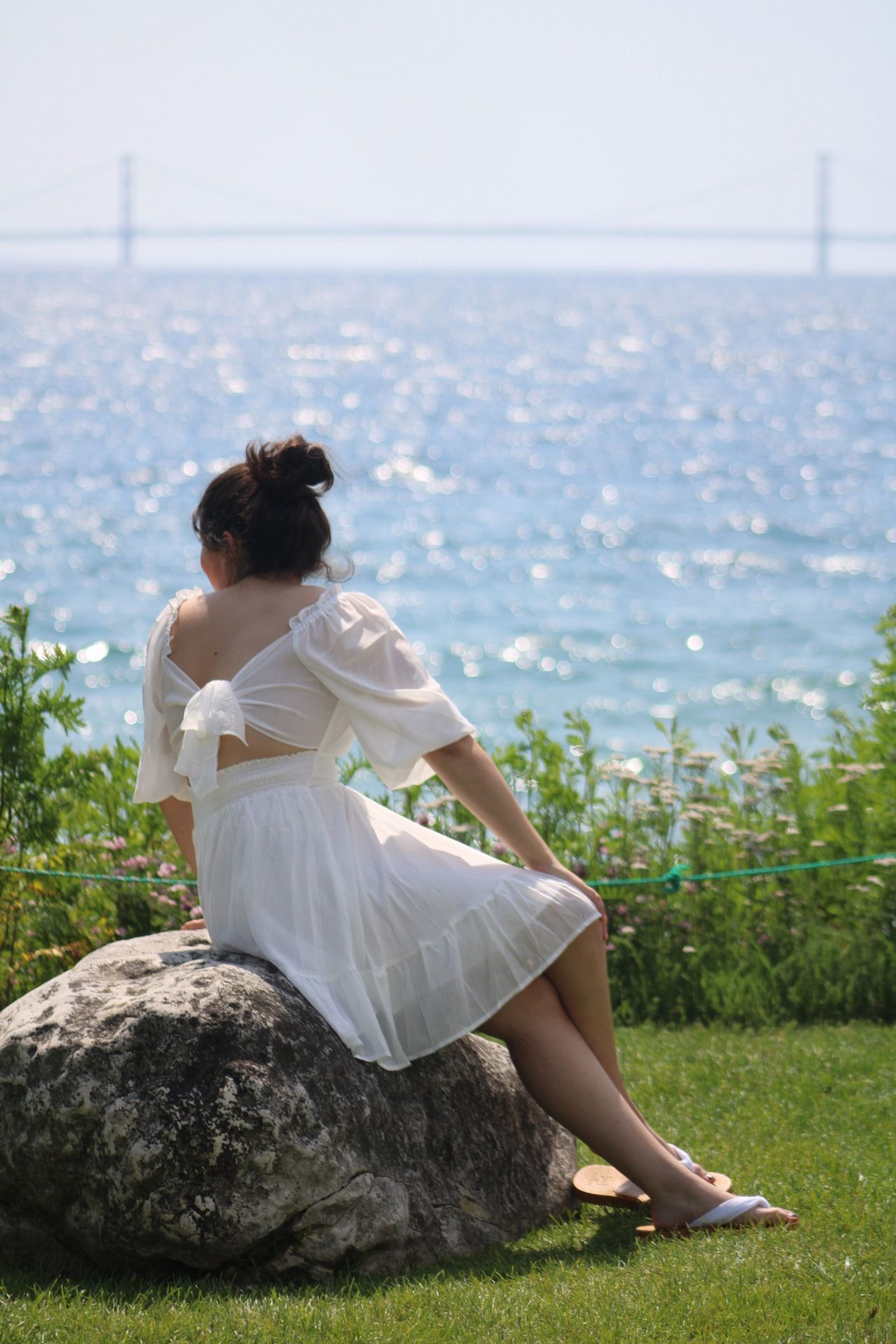 Woman in white dress sitting on rock overlooking sparkling blue water with Mackinac bridge in background on sunny day.