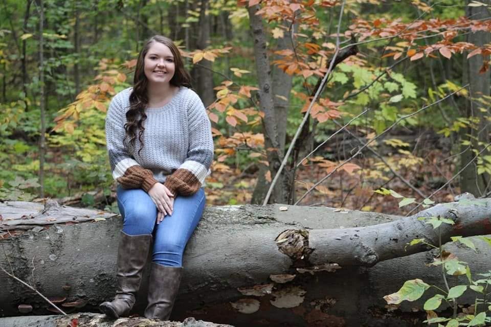 Young woman sitting on a fallen log in an autumn forest, wearing a gray sweater, jeans, and brown boots.