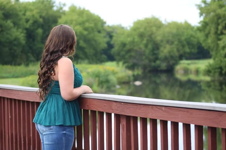 Young woman with curly brown hair in teal top and jeans stands on wooden bridge overlooking a serene green pond.