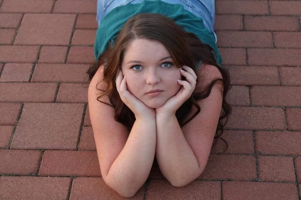 Young woman with brown hair lying on brick pavement, resting chin on hands, looking up thoughtfully.