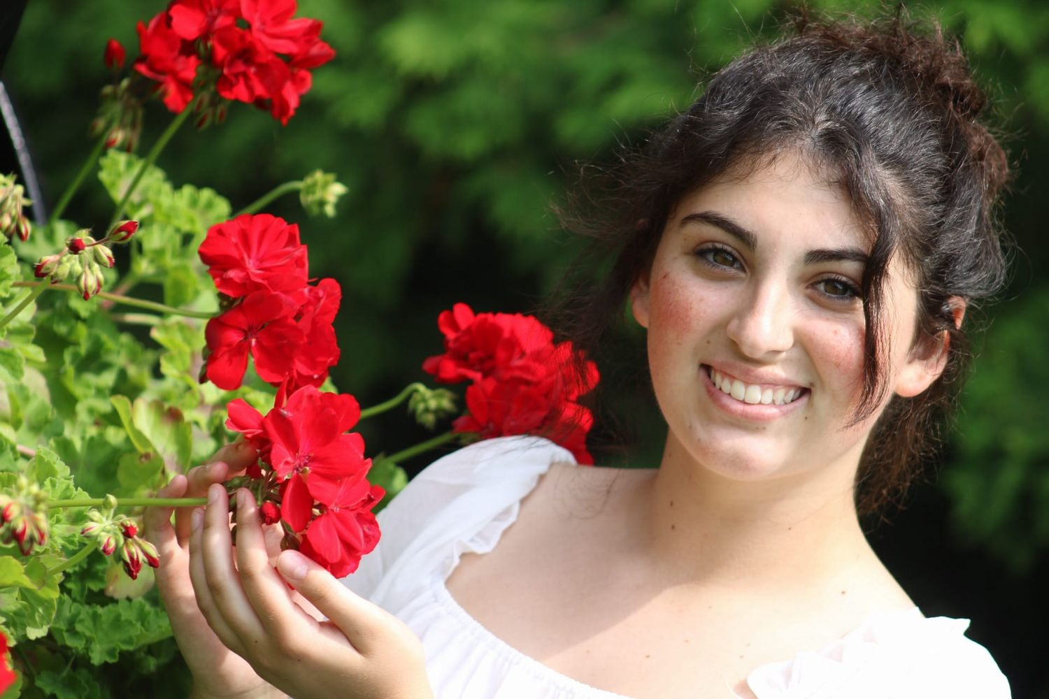 Young woman in white dress smiling beside vibrant red geranium flowers in a lush green garden.