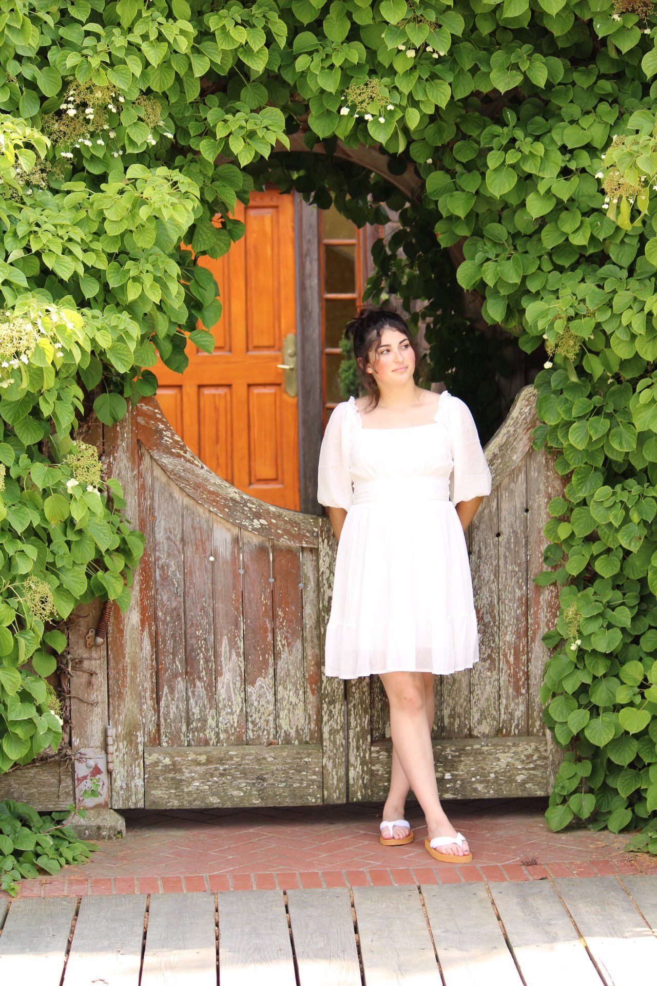 Young woman in white dress standing in rustic wooden garden gate framed by lush green ivy.