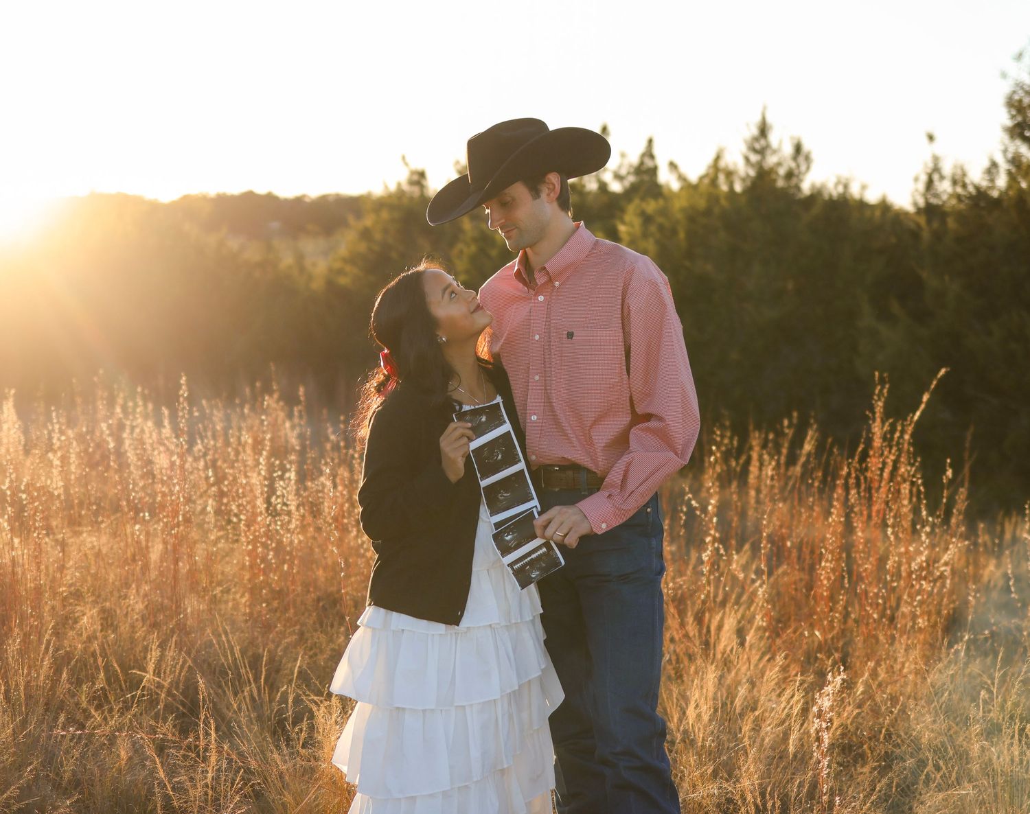 A romantic couple in western attire embracing in a golden sunlit field during sunset.