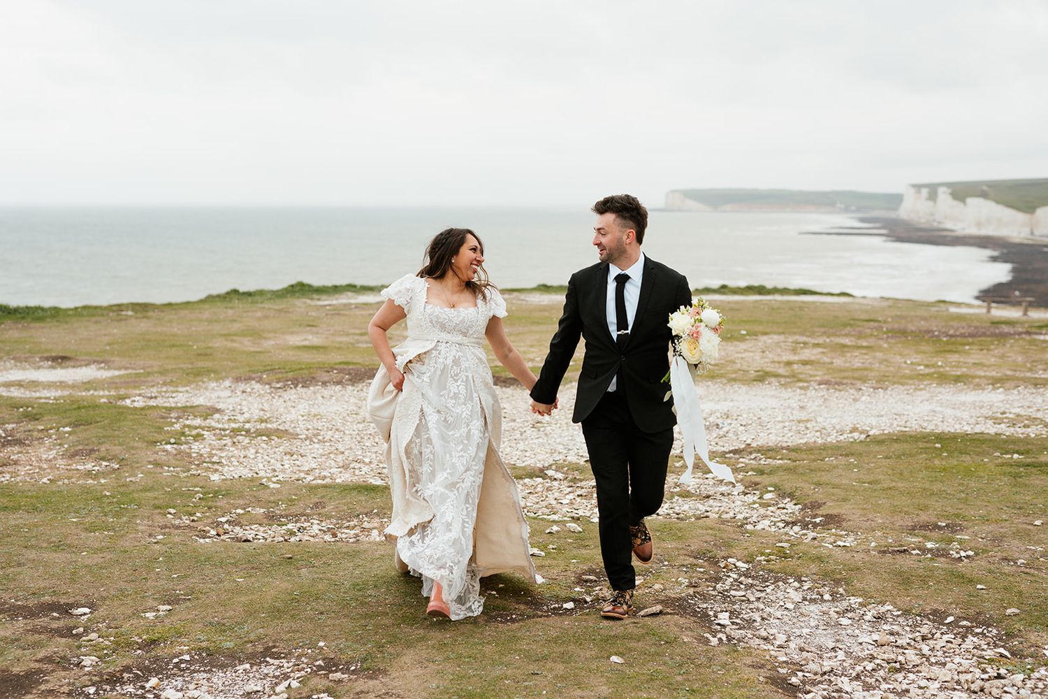 A couple in wedding attire poses dramatically against white cliffs at a scenic coastal location.