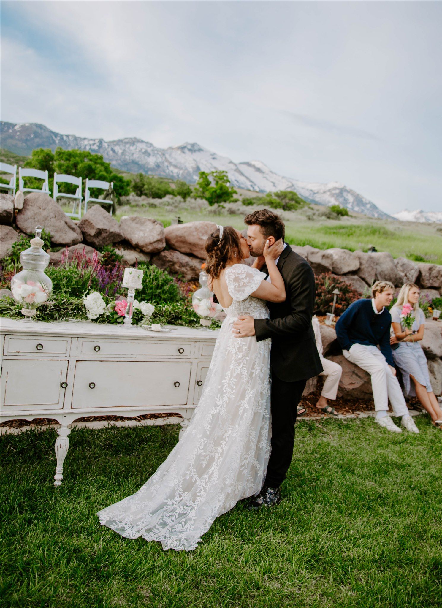 Wedding couple shares romantic moment on grassy hillside with mountain backdrop.