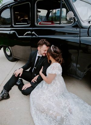 Newlyweds pose romantically beside a vintage black car on their wedding day.