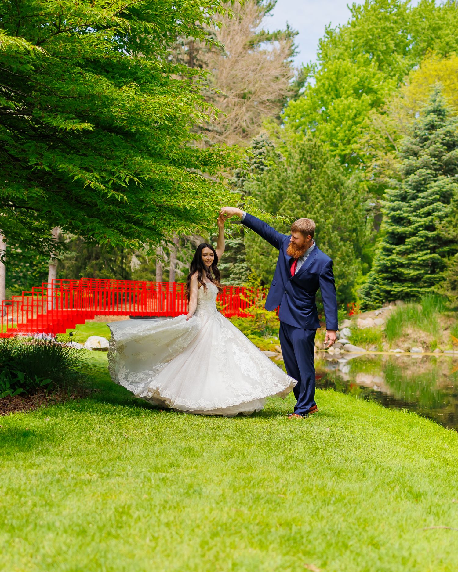 Couple dancing on green lawn with red bridge backdrop as bride's white dress twirls.