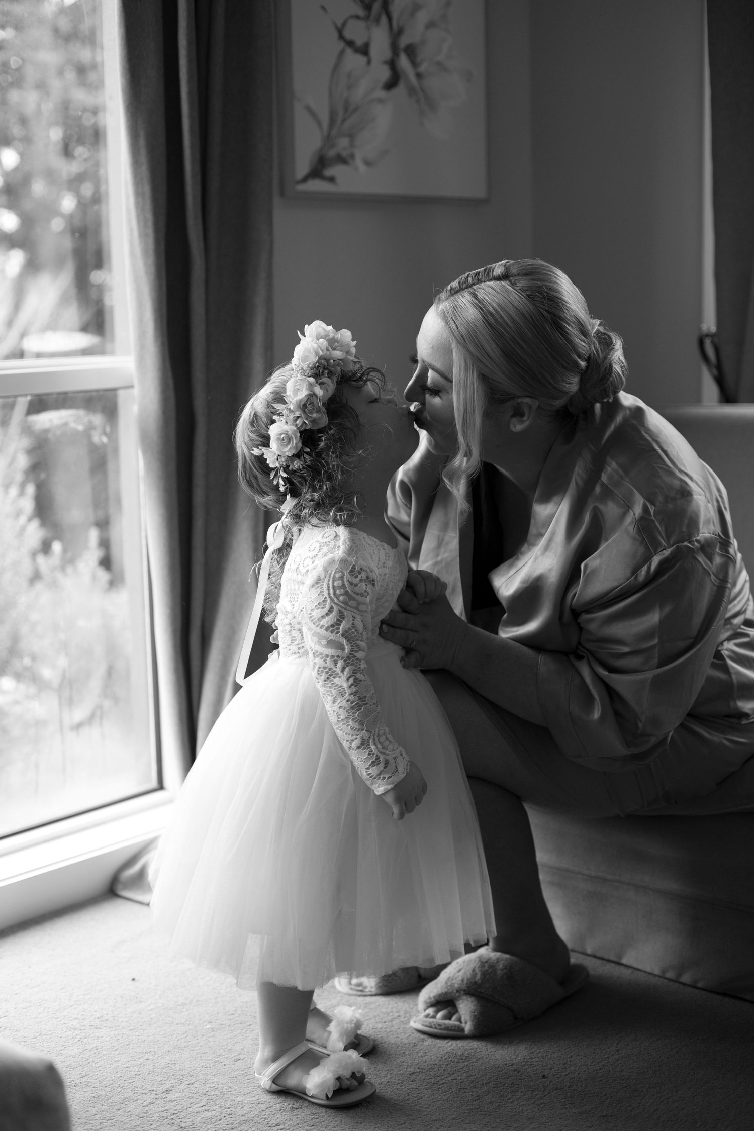 A tender black and white moment between a child in a white lace dress and adult near a window.